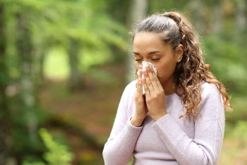 Woman blowing nose w tissue in nature 95325288 YI Woman experiencing seasonal airborne allergy symptoms from pollen