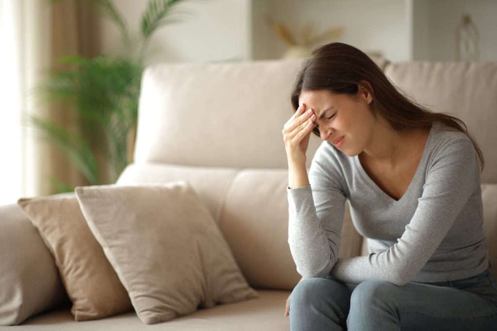 Woman w migraine on couch at home 98783944 YI Woman experiencing a severe migraine headache with forehead pain and light sensitivity while sitting on a couch.